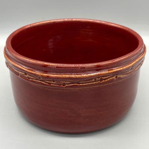 May include: A deep red ceramic bowl with a decorative band around the top. The band is a lighter brown color and has a woven design.
