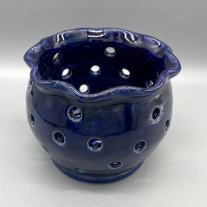 May include: A dark blue ceramic pot with a ruffled rim and numerous circular holes. The pot has a glossy finish and a rounded shape, suitable for holding small plants or decorative items.