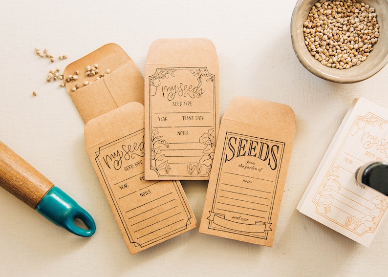 May include: Four brown paper seed packets with labels that say "my seeds" and "seeds from the garden of". The labels have space to write the year, seed type, plant date, and notes. There are also some seeds scattered on the table.
