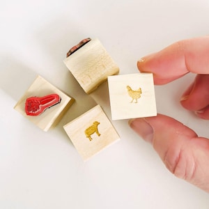 May include: Wooden stamps with farm animal designs. The stamps include a red pepper, a chicken, a sheep, and a stamp with a brown design. The stamps are on a white surface.