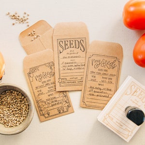 May include: Four brown paper seed packets with handwritten labels, each with a different type of seed. The labels include the seed type, year, plant date, and notes. The packets are on a white surface with a wooden stamp that says "Seeds" and two red tomatoes.