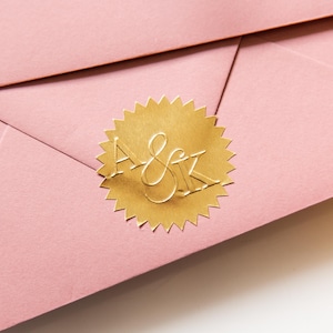 Pode incluir: Um envelope rosa com um adesivo de folha de ouro que diz "A & K" em letra cursiva.