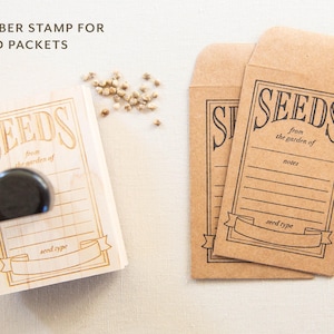 May include: A wooden rubber stamp for seed packets with the word "SEEDS" in a decorative font. Two brown seed packets are also visible, each with the word "SEEDS" printed on them. The text "from the garden of" is also visible.