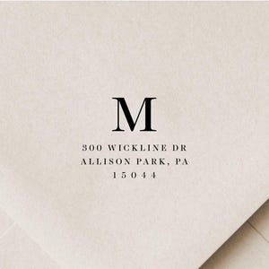 Puede incluir: Sobre color crema con una gran letra negra "M" en la parte superior. Debajo, la dirección "300 Wickline Dr, Allison Park, PA 15044" está impresa en una fuente sencilla y elegante. El sobre tiene una textura sutil.