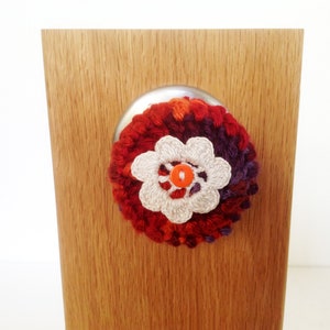 May include: A crocheted flower with a white center and red, orange, and purple petals. The flower is attached to a wooden surface.