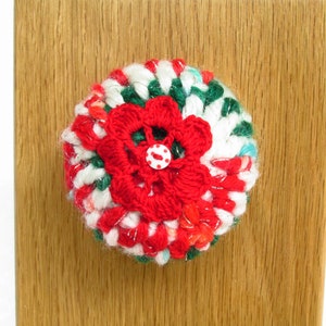 May include: A red, white, and green crocheted knob with a red flower and a white button in the center. The knob is attached to a wooden surface.