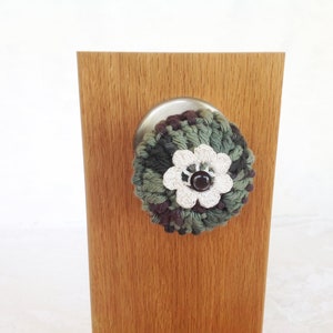 May include: A crocheted door knob cover in a camouflage pattern with a white flower and a brown button in the center. The cover is attached to a wooden door knob.