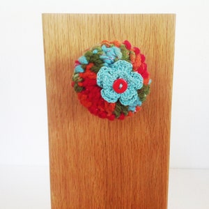 May include: A colorful crocheted flower with a red button center, attached to a wooden surface.