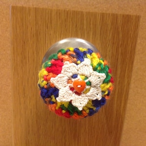 May include: A colorful crocheted doorknob cover with a white flower in the center and an orange button.
