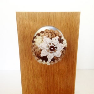 May include: A crocheted flower with brown, beige, and white yarn, attached to a wooden surface.