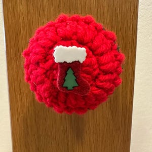May include: A circular red crocheted ornament featuring a central white and red stocking embellishment. The stocking has a white cuff and a small green Christmas tree icon centered on the front. The textured yarn creates a dense, rounded shape.
