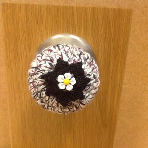 May include: A crocheted door knob cover with a white and black flower design. The flower has a yellow center.