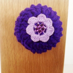 May include: A purple crocheted flower with a light purple crocheted flower in the center. The center flower has purple sequins.