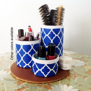 May include: Three blue and white patterned cylindrical containers with a white rim, holding various makeup and hair care items. The containers are on a brown circular base, sitting on a floral patterned surface.