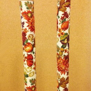 May include: Two white cylindrical chair leg covers with a floral pattern in shades of red, orange, yellow, green, and brown. The pattern features leaves, flowers, and fruit.