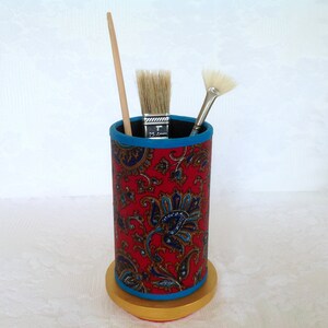 May include: Red and blue patterned fabric pencil holder with a gold base. The holder has three paint brushes inside.