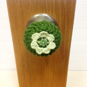 May include: A green and white crocheted flower with a green button in the center, attached to a silver door knob.