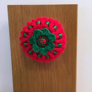 May include: A red and green crocheted flower with a button in the center. The flower is attached to a wooden surface.