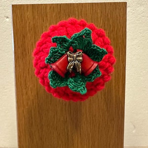 May include: A circular red crocheted ornament featuring a cluster of green crocheted holly leaves in the center. Two small red bells with a metallic gold-toned bow accent are attached to the middle of the green leaves, creating a festive design.