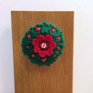 May include: A circular dark green crocheted ornament featuring a red crocheted flower at the center, accented with a small white button with red dots. The green base is woven with thin red ribbon strips throughout the textured yarn surface.
