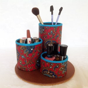 May include: A red and blue paisley patterned makeup organizer with three compartments. The organizer has a round base and holds makeup brushes, lipstick, and nail polish.