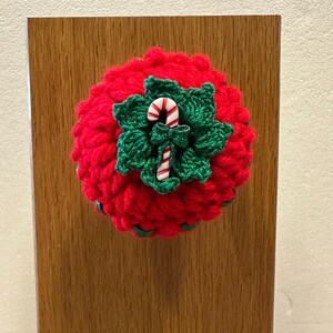 May include: A round, red crocheted ornament featuring a central cluster of dark green crocheted leaves. A small, white and red striped candy cane charm with a tiny green bow is attached to the center of the green leaves, creating a festive design.