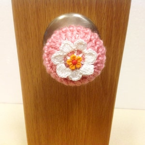 May include: A pink and white crocheted flower with an orange button center covers a door knob.