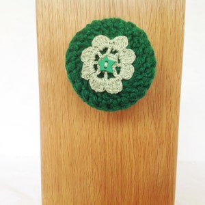 May include: A green crocheted flower with a green star button in the center. The flower is attached to a wooden surface.