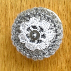May include: A gray crocheted flower-shaped knob cover with a light gray flower center and a dark gray button. The knob cover is set against a wooden surface.