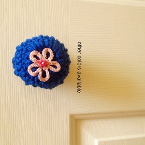 May include: A blue crocheted door knob with a pink flower on top. "other colors available" is written on the door.