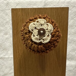 May include: A round, textured, brown crocheted piece featuring a cream-colored crocheted flower centered with a small, dark brown four-hole button. The item is mounted on a vertical, light-colored wooden board with a visible wood grain pattern.