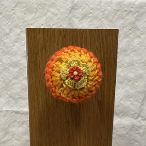 May include: A round, crocheted knob cover featuring a gradient of orange, yellow, and red yarn. The center is adorned with a small, layered, light yellow crocheted flower and a tiny, red, five-petaled plastic flower button with a yellow center.