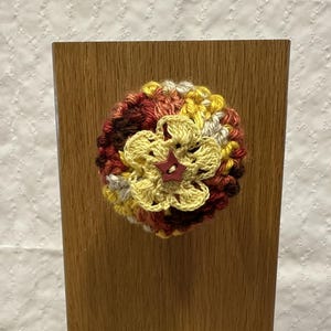May include: A round, multi-colored crochet flower applique featuring layers of yellow, red, and brown yarn. The center of the flower is adorned with a small, dark red, five-pointed star-shaped button, creating a textured, layered floral design.