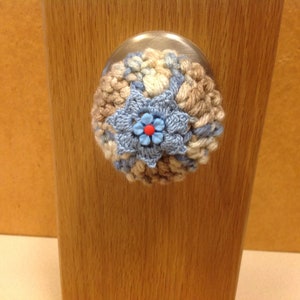 May include: A crocheted door knob cover in shades of brown, beige, and blue with a blue flower in the center. The flower has a red center.