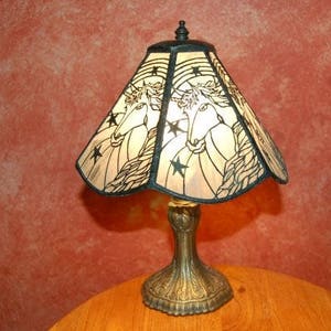 May include: A table lamp with a stained glass shade featuring a unicorn design. The shade is a light brown color with black outlines and stars. The base of the lamp is a dark bronze color.