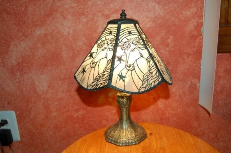 May include: A table lamp with a stained glass shade featuring a unicorn design. The shade is a light brown color with black outlines and stars. The base of the lamp is a dark bronze color.