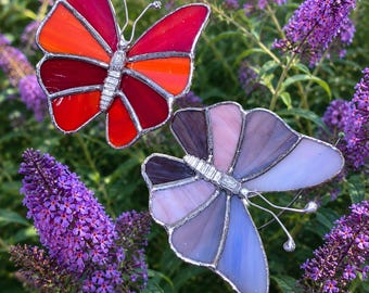 May include: Two stained glass butterfly garden stakes. One butterfly has red and orange wings, the other has purple and blue wings. Both have silver metal bodies and antennae. The butterflies are set among purple flowers and green foliage.