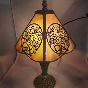 May include: A vintage-style table lamp with a brown metal base and a stained glass shade featuring a floral design. The shade is a warm amber color with a scalloped edge.