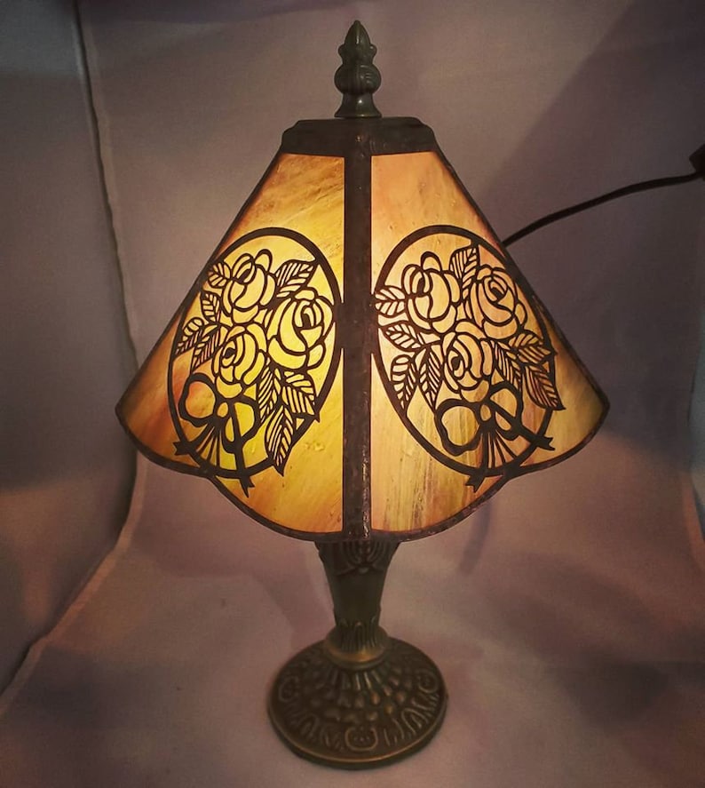 May include: A vintage-style table lamp with a brown metal base and a stained glass shade featuring a floral design. The shade is a warm amber color with a scalloped edge.