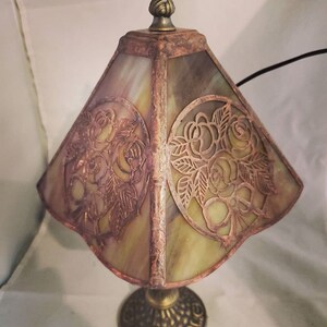 May include: A small, antique-style table lamp with a stained glass shade featuring a floral design. The shade is a pale green with a copper-toned floral pattern. The base is a dark bronze with a decorative design.