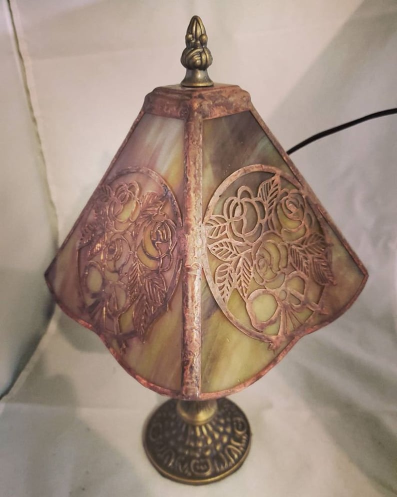 May include: A small, antique-style table lamp with a stained glass shade featuring a floral design. The shade is a pale green with a copper-toned floral pattern. The base is a dark bronze with a decorative design.