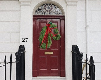 May include: A red front door with a green wreath and red ribbon. The door is part of a white building with the number 27 above the steps. A black wrought iron fence and steps lead up to the door.
