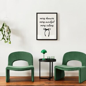 May include: Two green velvet chairs with a black metal side table and a green lamp in between. The wall behind the chairs has a black framed print with the text "very demure very mindful very cutesy" and a black bow.
