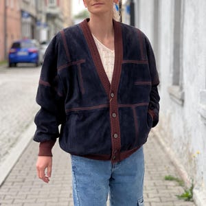 May include: A navy blue suede jacket with brown trim and button closure. The jacket has a V-neck and is paired with light blue jeans. The jacket features a ribbed brown cuff and trim. The outfit is worn outdoors.