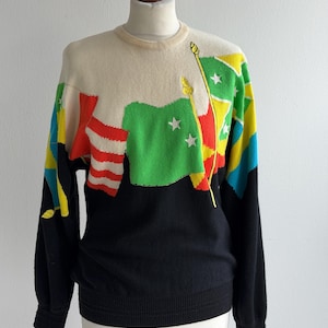 May include: A vintage sweater with a distinctive flag design. The upper section is cream, showcasing colourful flag graphics in red, green, yellow, and blue. The lower half and sleeves are black. The sweater is displayed on a mannequin.