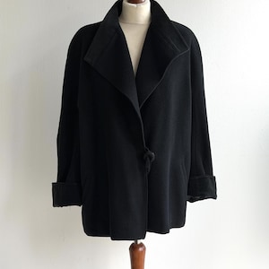 Pu&ograve; includere: Un cappotto nero con ampio colletto e maniche lunghe, esposto su un manichino di legno. Il cappotto presenta una chiusura con un solo bottone e polsini risvoltati. La lunghezza del cappotto arriva a met&agrave; coscia.