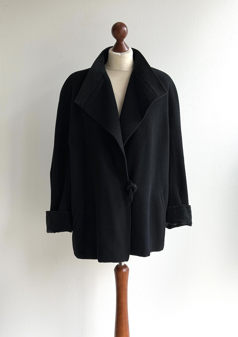 Pu&ograve; includere: Un cappotto nero con ampio colletto e maniche lunghe, esposto su un manichino di legno. Il cappotto presenta una chiusura con un solo bottone e polsini risvoltati. La lunghezza del cappotto arriva a met&agrave; coscia.