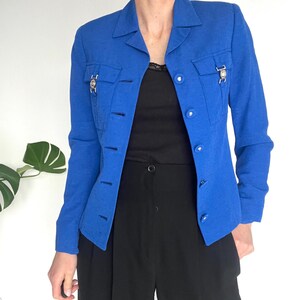 May include: A bright blue blazer with a collared neckline, long sleeves, and button closures. The blazer has two flap pockets with silver accents and a textured fabric.