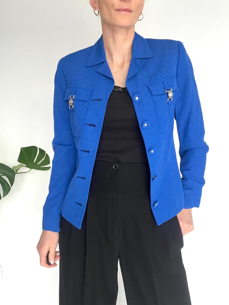 May include: A bright blue blazer with a collared neckline, long sleeves, and button closures. The blazer has two flap pockets with silver accents and a textured fabric.