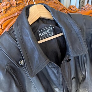 May include: A black leather jacket with a notched collar and button closures. The jacket is hanging on a wooden hanger. The label reads "ROVEX Leather Fashions".